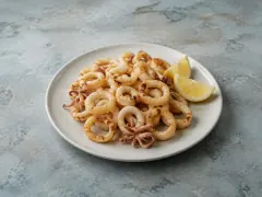 A plate of lightly battered fried squid rings and tentacles, pale gold, with two lemon wedges on a pale blue-grey table.