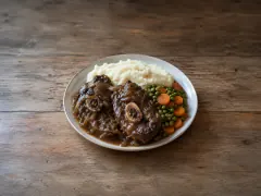 Two braised mutton chops in dark onion gravy on a plate with a mound of mashed potato and peas and carrots.