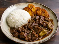 Dark roucou-tinted game fricassee with tender meat cubes and peppers, plated with white rice and fried plantain.