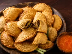 A pile of small golden half-moon fried pastels with crimped edges, one opened to show spiced beef, beside red pepper sauce.