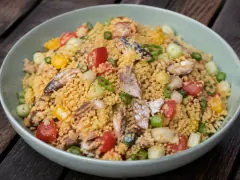 Golden toasted cassava grains tossed with cucumber, tomato, red onion, peppers, herbs and flakes of smoked fish in a pale bowl.