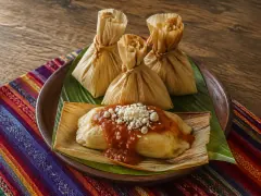 Three corn-husk parcels tied at the top like little sacks on a green leaf, beside one unwrapped pale yellow masa dumpling topped with red tomato sauce and crumbled white cheese, on a striped woven cloth.