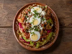 A painted terracotta plate holding stacked crisp corn tostadas topped with minced beef, red salsa, shredded lettuce, magenta pickled vegetables, three slices of hard-boiled egg, crumbled white cheese and chopped herbs.