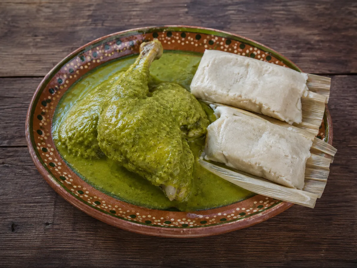 A painted terracotta plate holding a chicken leg quarter blanketed in vivid jade-green sauce, beside two pale corn tamalitos still in their husks, on a dark wooden table.