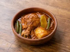 A round terracotta bowl of thick brick-red stew holding a chicken leg on the bone, a wedge of pale green squash, whole green beans and a chunk of potato, on a dark wooden table.
