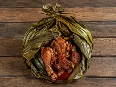 A large green leaf parcel tied at the top and folded open like petals, holding pieces of chicken on the bone and dark braised meat in a thick deep-red sauce, on a wooden table.