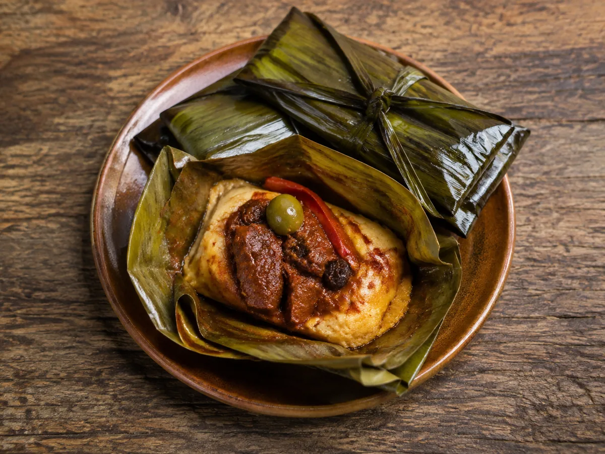 A round terracotta plate holding an opened banana-leaf parcel of pale yellow masa topped with dark red sauce, a green olive and a strip of red pepper, with a second tied parcel behind it on a wooden table.
