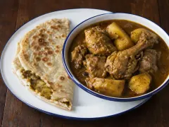 A bowl of golden chicken curry with potato pieces next to a folded dhalpuri roti torn open to show its yellow split-pea filling.