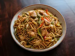 A bowl of Guyanese chowmein: brown-glazed noodles tossed with chicken strips, red pepper, green beans and spring onion.