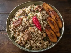 A bowl of coconut cook-up rice with black-eyed peas, chunks of beef and shrimp, a whole red pepper on top and fried plantain slices at the side.