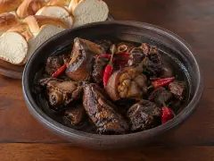 Dark, glossy pepperpot of beef and pork in black cassareep gravy with whole red peppers, in a black bowl beside a plait of soft white bread.
