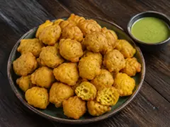 A heap of golden fried pholourie split-pea balls on a plate with a small bowl of green mango sour beside them.