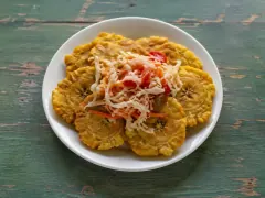 A white plate of flattened golden fried plantain discs with jagged edges, topped with a mound of shredded white cabbage and red pepper slaw, on a weathered green wooden table.