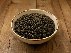 A rustic cream-rimmed bowl heaped with glistening jet-black rice studded with bright green peas, on a dark wooden table.