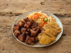 A speckled plate divided between deep mahogany fried pork cubes, a pile of shredded cabbage and carrot slaw topped with sliced red and green chilli, and flattened golden fried plantains.