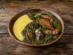 A terracotta bowl half filled with smooth golden-yellow cornmeal and half with a dark green leafy stew holding orange crab claws and chunks of pork, on a dark wooden table.