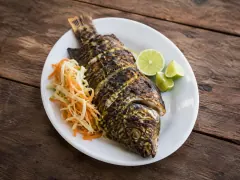 A whole charred grilled fish with diagonal score marks across its skin on a white oval plate, with shredded cabbage and carrot slaw and three lime wedges beside it.