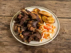A green-rimmed plate piled with dark bronze bone-in fried goat pieces, beside golden fried plantain slices and a heap of shredded cabbage and carrot slaw.
