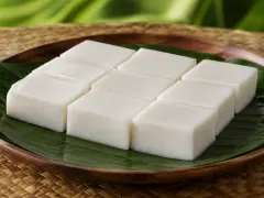 Nine porcelain-white squares of coconut haupia arranged on a green ti leaf on a wooden plate.