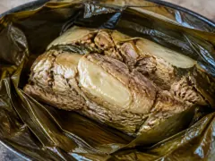 An unwrapped lau lau bundle on dark glossy leaves, with tender pork and a piece of pale fish inside collapsed taro greens.