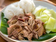 A plate of shredded kalua pork on a green leaf with two scoops of white rice and steamed cabbage wedges.