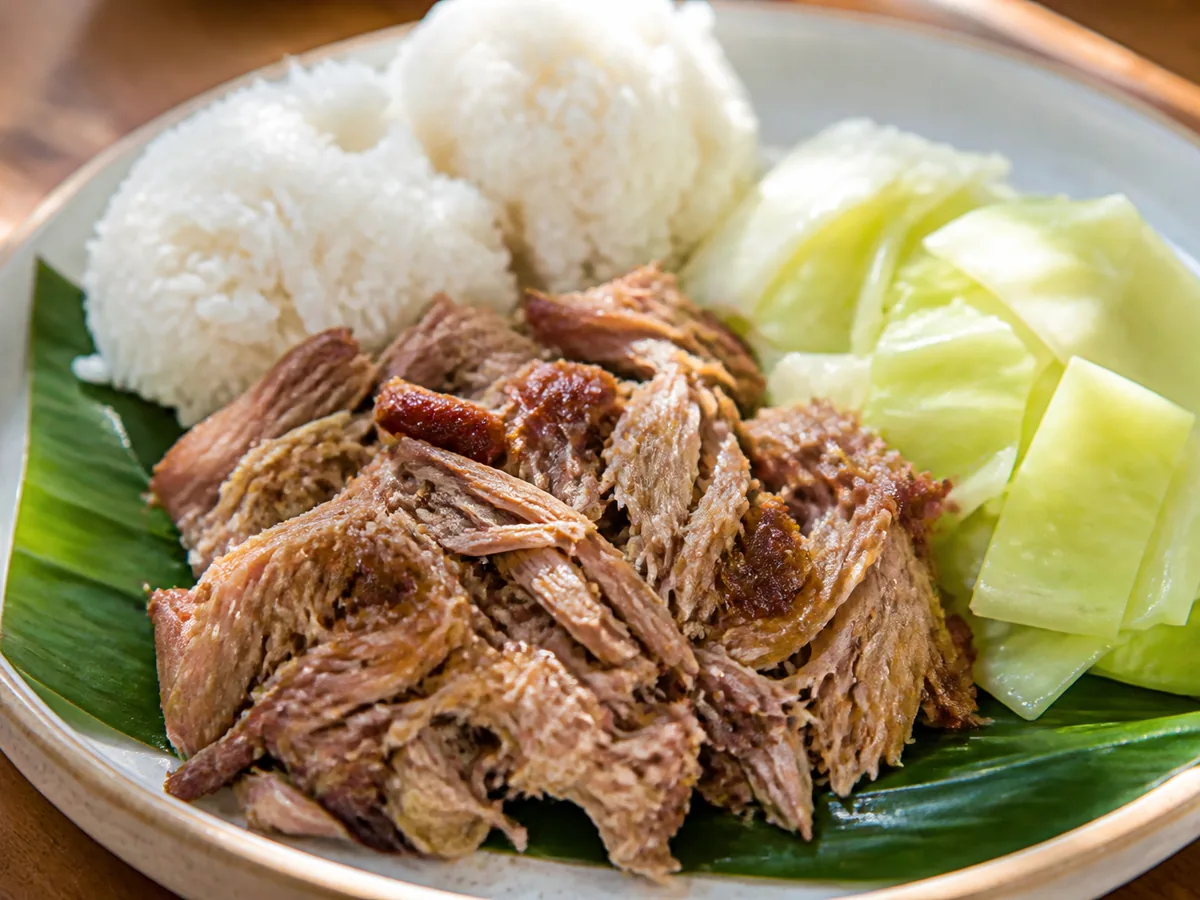 A plate of shredded kalua pork on a green leaf with two scoops of white rice and steamed cabbage wedges.