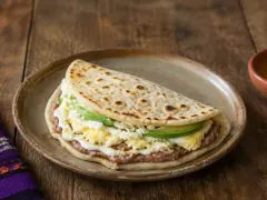 A large folded flour tortilla with pale golden blisters on a stoneware plate, its open side showing dark refried beans, white cream, scrambled egg, crumbled cheese and green avocado slices.