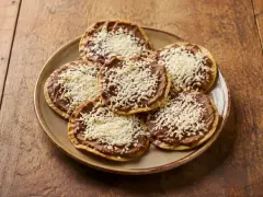 Six round fried corn tortillas on a stoneware plate, each spread with dark refried beans and heaped with finely grated white cheese.