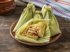 Three pale green corn-husk parcels tied at the ends on a terracotta plate, with one opened to show a yellow corn custard filled with shredded meat and red pepper.