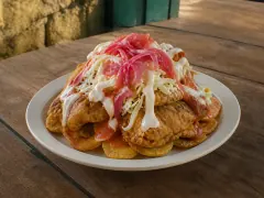 A mounded plate of golden fried plantain rounds and fried chicken pieces buried under shredded cabbage, pink pickled onions, red sauce and white creamy drizzle.