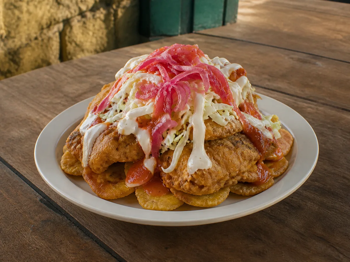 A mounded plate of golden fried plantain rounds and fried chicken pieces buried under shredded cabbage, pink pickled onions, red sauce and white creamy drizzle.