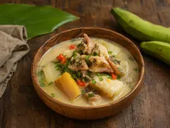 A carved wooden bowl of creamy pale coconut broth with pieces of white conch, yellow plantain, cassava and red pepper, scattered with chopped green herbs, beside green plantains on a wooden table.