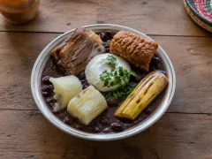 A rimmed bowl of dark reddish-brown bean soup holding whole beans, a piece of pork belly, a golden crackling, chunks of white cassava, a fried plantain and a spoonful of cream topped with cilantro.