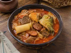 A dark bowl of orange-red stew holding chunks of beef, sliced sausage, cabbage, cassava and yellow plantain, set in a woven basket on a wooden table.