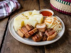 A rimmed plate of white boiled cassava chunks beside thick slices of fried pork belly, a pile of shredded cabbage and carrot slaw, and a small dish of red sauce.
