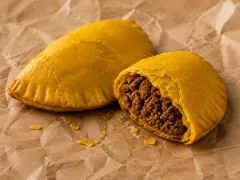 Golden-yellow half-moon beef patties with fork-crimped edges on a wire rack, one broken open to show the dark spiced beef filling inside flaky pastry.