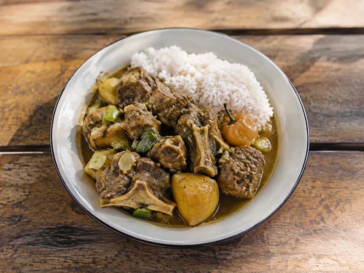 Chunks of bone-in goat and soft potato in a deep ochre curry gravy, served beside white rice with a scattering of sliced scallion.