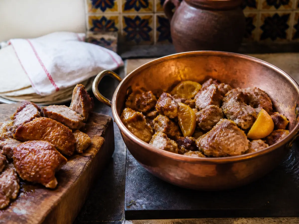 A copper pan of browned pork chunks with cooked orange pieces tucked among them, beside a wooden board of crisped pork and a cloth-wrapped stack of tortillas, against patterned tile.