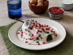 A single stuffed poblano with its stem still on, blanketed in ivory walnut sauce and scattered with pomegranate seeds and parsley, on a pale plate over a green napkin, with bowls of walnuts, pomegranate arils and cream behind.