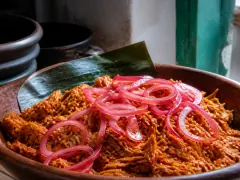 A wide clay bowl of orange-red shredded pork piled against a banana leaf, with bright pink pickled onion rings heaped across the middle.