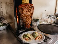 A trompo of stacked adobo-red pork on a vertical broiler, crowned with a whole pineapple, beside a plate of two open tacos topped with diced onion, cilantro and pineapple on a steel taquería counter.