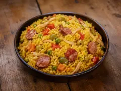 A dark bowl of golden-yellow rice studded with sliced red sausage, shredded chicken, green olives, peas, diced carrot and red pepper, on a wooden table.