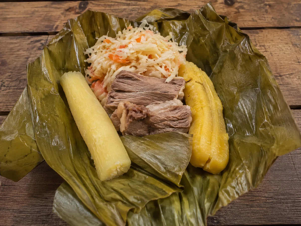 A green banana leaf spread with a length of pale cassava, a whole yellow plantain, slices of tender beef and a mound of shredded cabbage and carrot slaw, on a wooden table.
