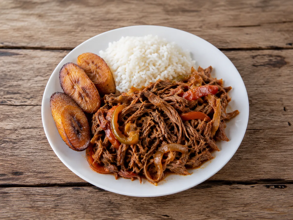 A white plate of long shredded beef in reddish-orange sauce with soft onion and pepper strips, beside a mound of white rice and three dark fried plantain slices.
