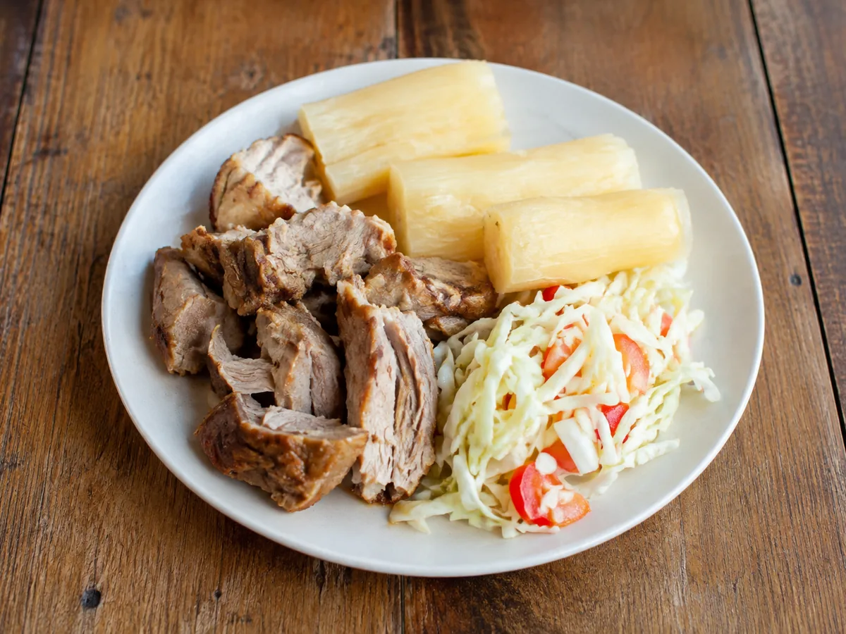 A white plate of browned pork pieces beside three lengths of pale boiled cassava and a heap of shredded cabbage slaw with diced red tomato, on a wooden table.