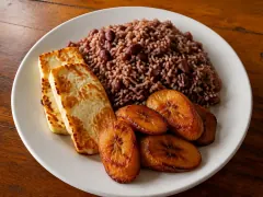 A white plate of deep reddish-brown rice studded with whole red beans, beside two golden-seared slabs of white cheese and four caramelised dark plantain slices.