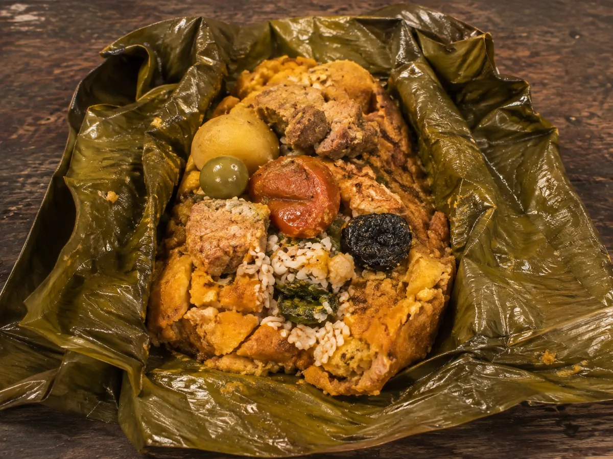 An opened green banana-leaf parcel on a dark table showing orange corn masa filled with chunks of pork, white rice, a boiled potato, a green olive, a tomato slice and a dark prune.