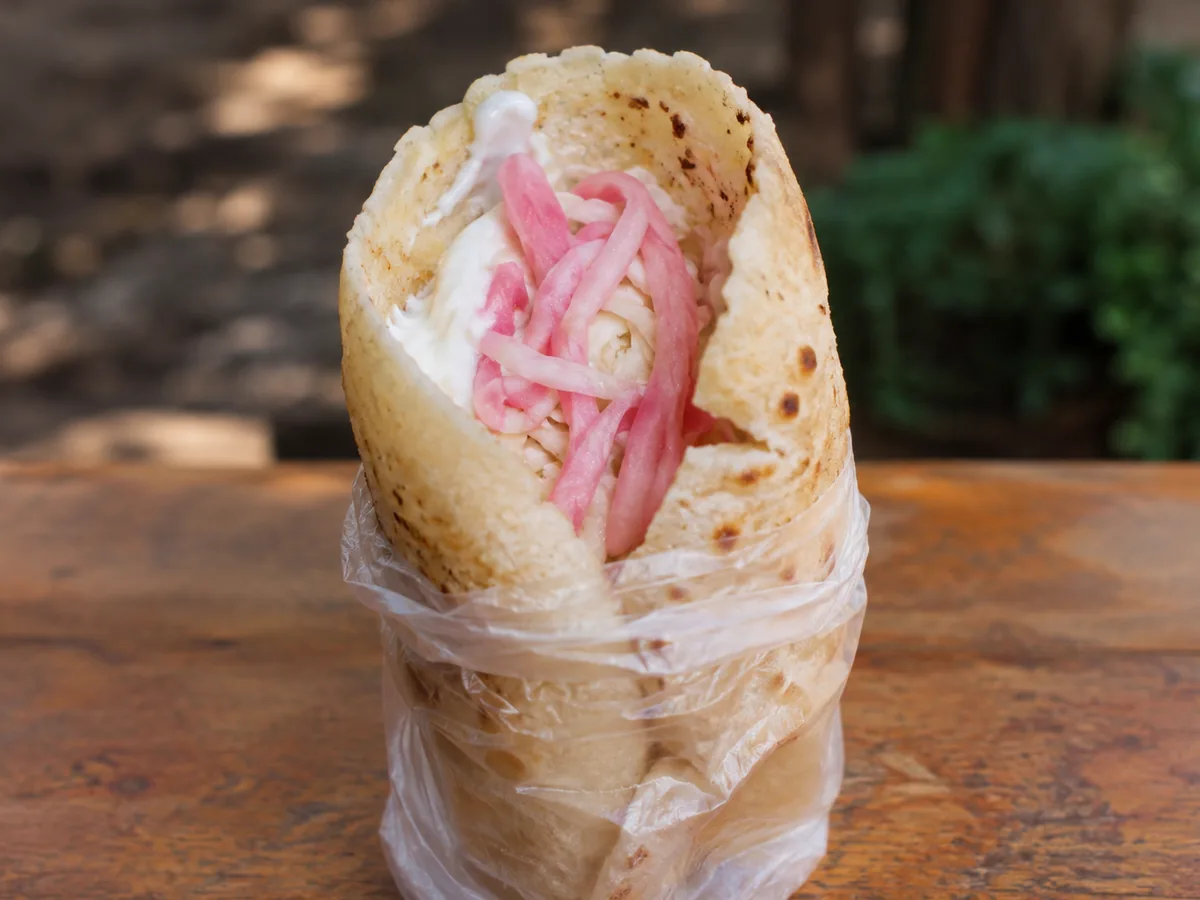 A rolled corn tortilla standing upright in a twisted plastic bag, its open top showing white cheese, thick cream and bright pink pickled onion strands.