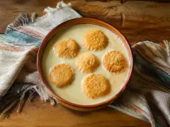 A terracotta bowl of pale golden creamy soup with six round golden-brown fried cheese fritters floating on the surface, on a striped woven cloth.