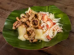 A large green banana leaf holding pale boiled cassava chunks, golden curled pork cracklings and a pile of shredded cabbage with diced tomato, on a wooden table.